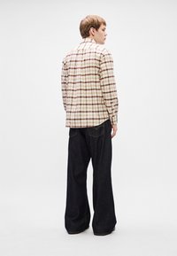 Camicia a scacchi nei toni del beige, marrone e rosso, abbinata a jeans in denim scuro di taglio largo. La camicia presenta un colletto con bottoni e maniche lunghe.