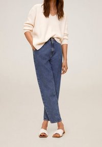 Un pull en tricot beige clair, un jean en denim bleu taille haute et des sandales à glissière blanches. Le jean a une coupe décontractée avec des chevilles retroussées.