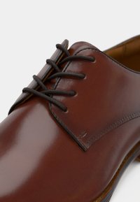 Chaussure en cuir marron à finition brillante, avec des lacets brun foncé, un bout rond et une couture discrète le long du bord supérieur.