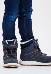 Marineblauwe winterlaarzen met rubberen zolen, verstelbare riemen en textiele stof. Witte nepbontvoering aan de bovenkant met oranje accenten.