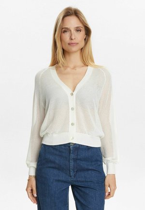 Weißer Strickcardigan mit V-Ausschnitt, langen Ärmeln und Knopfleiste. Besitzt ein offenes Netzdesign und einen gerippten Saum. Getragen mit blauen Jeans.