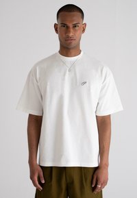 White cotton t-shirt with short sleeves, round neckline, and a small maroon logo on the chest. Worn with olive green pants.