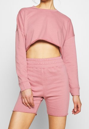 Person iført en lyserød cropped langærmet sweatshirt og matchende højttaljede shorts med elastik i taljen.