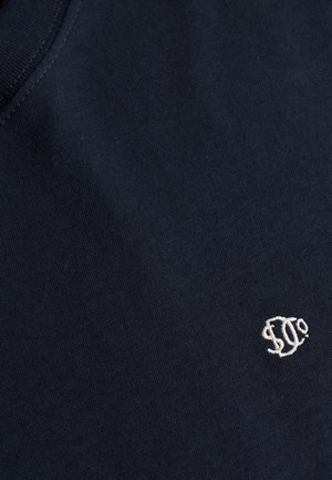 Tissu bleu foncé avec un petit logo brodé blanc composé de lettres stylisées près du bord, montrant la texture du tissu et les détails de la couture.