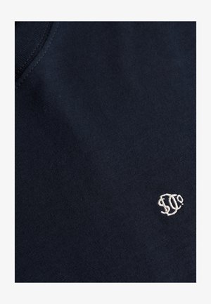 Donkerblauwe stof met een klein wit geborduurd logo met gestileerde letters nabij de rand, met zicht op de stofstructuur en stiksel details.