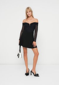Vestido preto off-the-shoulder com mangas longas transparentes, corpete justo e saia rodada. Usado com saltos pretos slingback e uma bolsa branca.