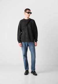 Sudadera negra con un patrón sutil, vaqueros azules, zapatos negros y gafas de sol rectangulares; el modelo posa contra un fondo claro y liso.