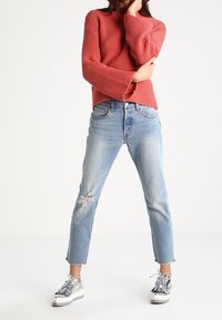 Frau in einem strukturierten roten Pullover, hellblauen, zerrissenen Jeans und glänzenden silbernen Plateauschuhen, die mit gespreizten Beinen vor weißem Hintergrund steht.