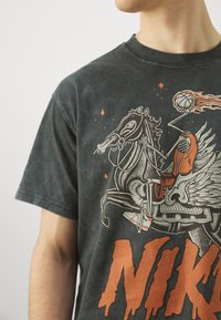 Schwarzes Baumwoll-T-Shirt mit einem Grafikdesign eines geflügelten Pferdes und eines Skeletts, mit orangefarbenen Akzenten und einem auffälligen „NIKE“-Text am unteren Rand.
