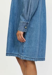 Rochie din denim de culoare albastru deschis, cu manșete cu nasturi, croială dreaptă și cusături subtile la tiv. Textură netedă și greutate medie.