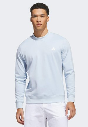 Mladý muž ve světle modré mikině s dlouhým rukávem a logem Adidas, v bílých kalhotách, stojící před jednoduchým světlým pozadím.