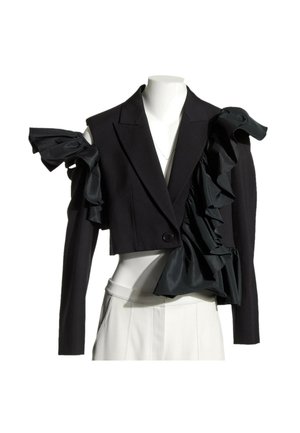 Blazer court noir avec des détails volantés exagérés sur une épaule et un revers, porté sur un pantalon blanc taille haute sur un mannequin.