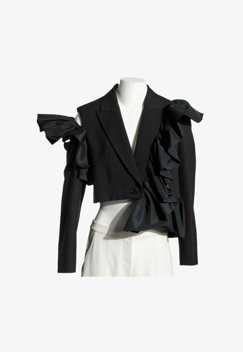Blazer court noir avec des détails volantés exagérés sur une épaule et un revers, porté sur un pantalon blanc taille haute sur un mannequin.