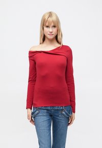 Červený top s dlouhým rukávem a odhalen�ými rameny s ozdobou z překroucené tkaniny u výstřihu, ve spojení s modrými džínami s kapsami.