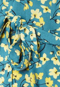 Tecido de vestido floral com fundo turquesa, flores amarelas e ramos pretos. Inclui um detalhe de cintura acomodada com uma textura suave.