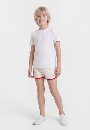 Szőke fiú áll és mosolyog, egyszerű fehér pólót, bézs rövidnadrágot piros szegéllyel, valamint fehér tornacipőt visel, fehér háttér előtt.