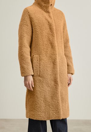 Long manteau teddy couleur camel avec col haut, poches latérales et fermeture frontale dissimulée porté sur un pantalon sombre.