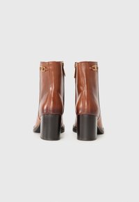 Bottines en cuir marron avec un talon bloc haut, fermeture zippée à l'arrière et accents en métal doré près de la cheville. Texture lisse.
