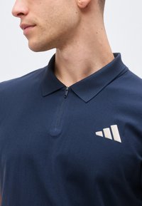 Homme portant un polo bleu marine avec une courte fermeture éclair et un logo triangulaire blanc sur la poitrine gauche, tourné vers la gauche.