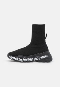Zapatilla tipo calcetín negra con una parte superior de punto texturizado, que presenta una suela negra gruesa y el logo en blanco de "versacejeans couture" en el lado.