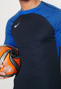 Marinblå sportskjorta med blårandiga ärmar, med en vit Nike-logotyp; personen håller en orange och svart fotboll.