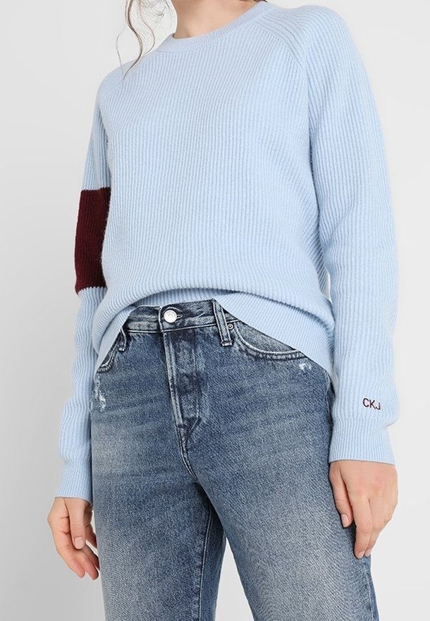 Light blue ribbed sweater with a maroon accent on the left sleeve, paired with distressed blue jeans. Features a round neckline and fitted cuffs.