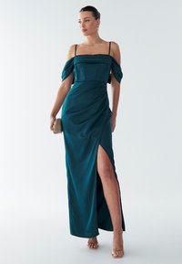 Teal satijnen jurk met off-shoulder mouwen en een zijsplit. Heeft een aansluitende corsage en een vloeiende roktextuur, aangevuld met strappy hakken.