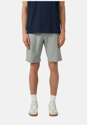 Mężczyzna ubrany w jasnoszorty do kolan, ciemnogranatową koszulę, białe długie skarpetki i białe sportowe buty stoi na jednolitym tle.