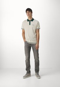 Polo shirt crema con colletto e rifiniture verdi; texture rigata; abbinato a jeans grigi e sneaker grigie chiare. Dettaglio delle stringhe blu.