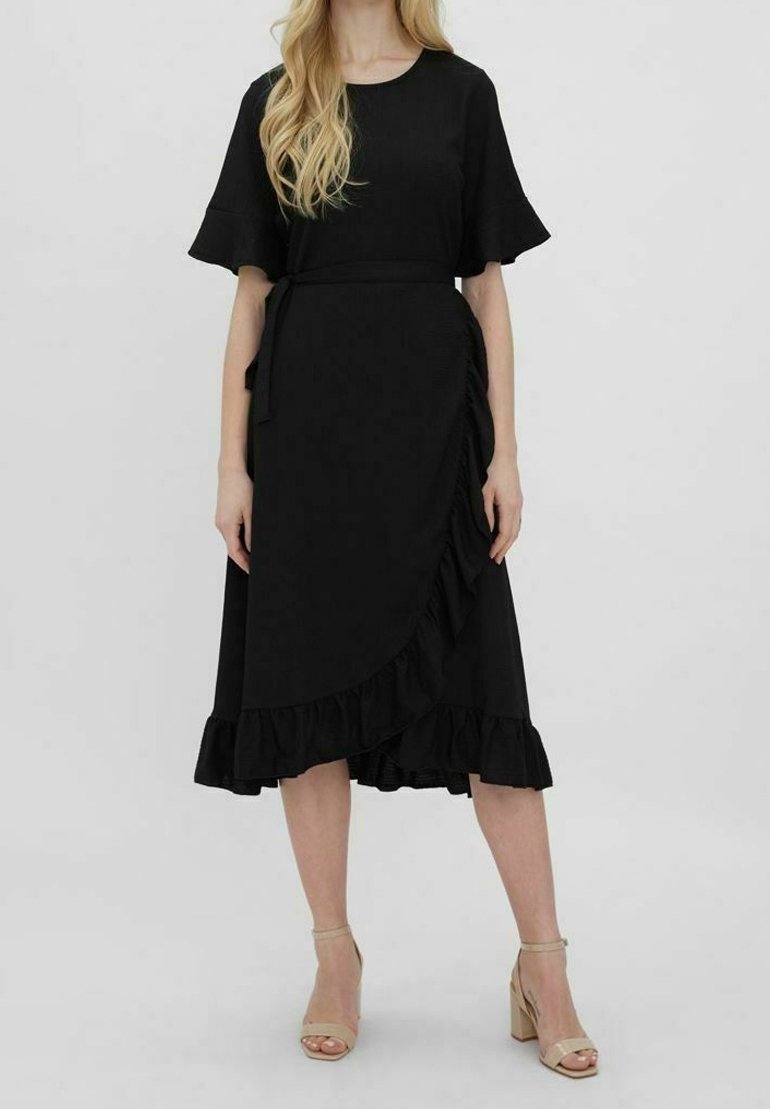 Femme portant une robe midi noire avec un ourlet et des manches à volants, nouée à la taille, associée à des sandales beiges à talons blocs avec bride à la cheville.