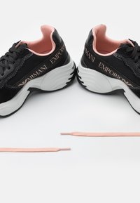 Svarta och rosa sneakers med en texturerad ovansida, tjock vit sula och logodetaljer på sidan. Inkluderar matchande rosa snörning.