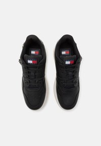 Black sneakers with a smooth leather upper, perforated details, textured mesh, and a white outsole. Features branding tags in red, white, and blue.