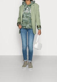 Personne portant un blazer vert sur un sweat à capuche camouflage GAP, un jean bleu, des baskets vertes, et tenant un sac à main matelassé blanc dans la main droite.