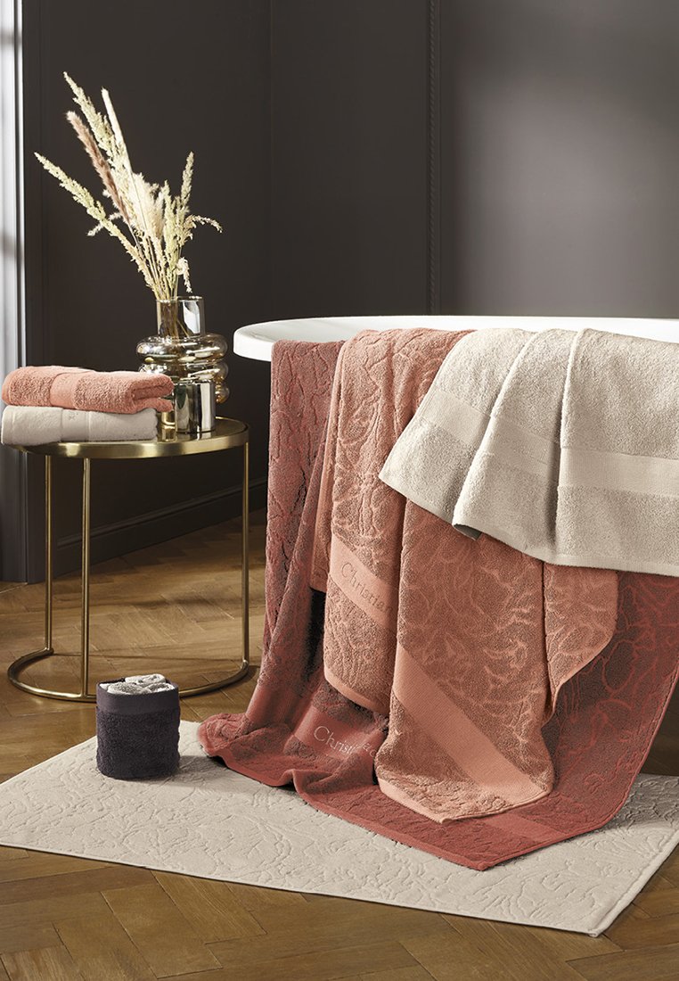 Asciugamani in varie tonalità di rosa, crema e carbone, con motivi floreali e texture. Un tappetino da bagno beige e un tavolino laterale metallizzato.