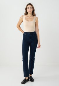 Donna in piedi contro uno sfondo neutro, che indossa un top sans maniche beige, jeans a vita alta blu scuro e mocassini neri.