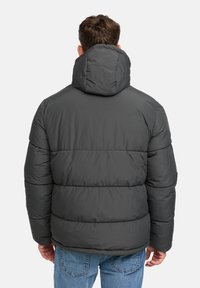 Man in een donkergrijze gewatteerde jas met capuchon en blauwe jeans, gezien vanaf de achterkant tegen een effen witte achtergrond.