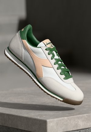 Biały sneaker z zielonymi sznurowadłami i wewnętrzną wyściółką, beżowe logo po bokach oraz brązowa podeszwa, pokazany w pozycji unoszącej się w powietrzu nad kamienną powierzchnią.