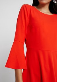 Robe rouge en tissu lisse avec un corsage ajusté et des manches évasées trois-quarts, dotée d'un col rond et d'un design minimaliste.