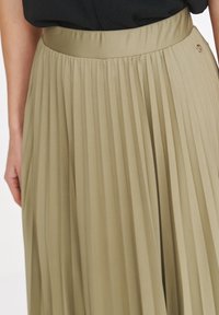 Plisowana spódnica w oliwkowej zieleni z gładkim pasem w talii i małym metalowym akcentem. Posiada pionowe fałdy tworzące fakturę.