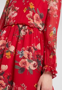 Robe fleurie rouge avec de longues manches transparentes, taille froncée et poignets froncés, sur un bras et une main d'une personne visibles.