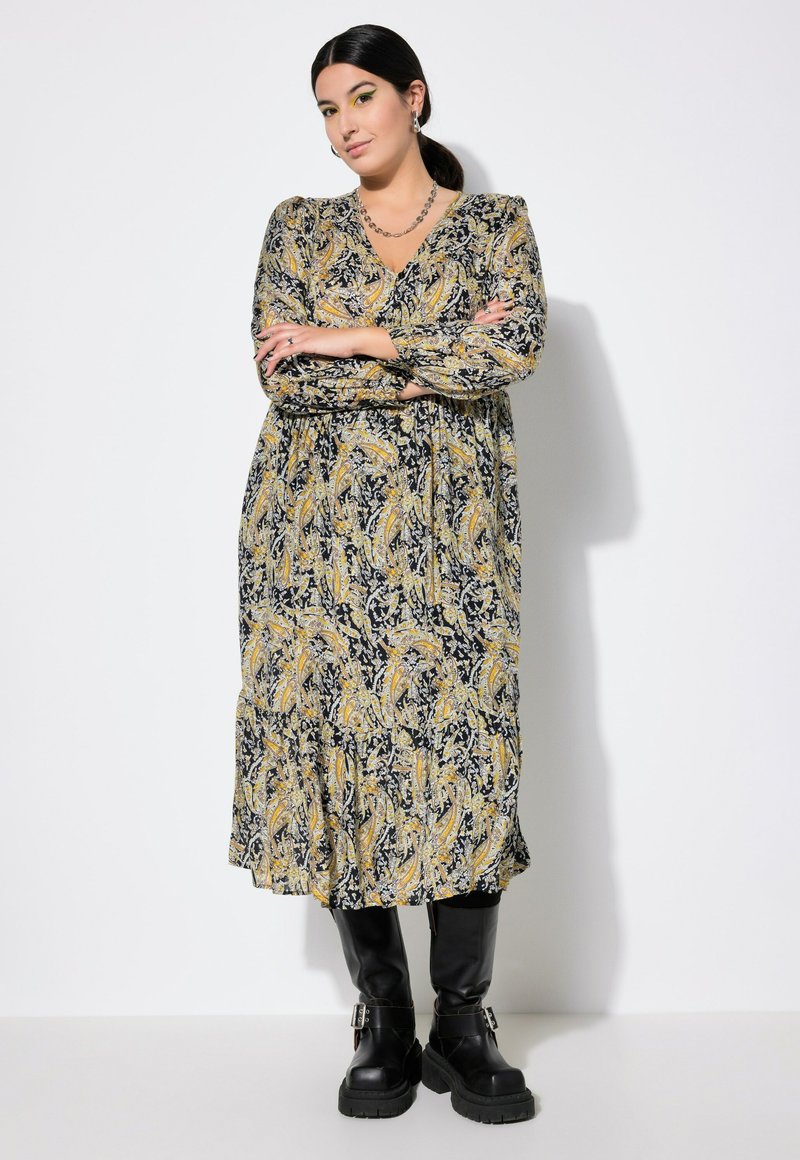 Robe longue à fleurs noire, jaune et blanche avec décolleté en V et manches longues, associée à des bottes noire à hauteur de genou avec des boucles.