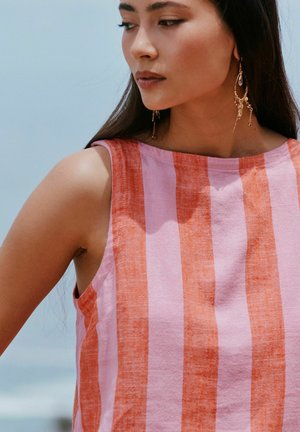 Pitkähiuksinen nainen, jolla on punaisen ja vaaleanpunaisen pystysuuntaiset raidat sisältävä hihaton paita ja kultaiset roikkuvat korvakorut, katsoo alas ja sivulle.