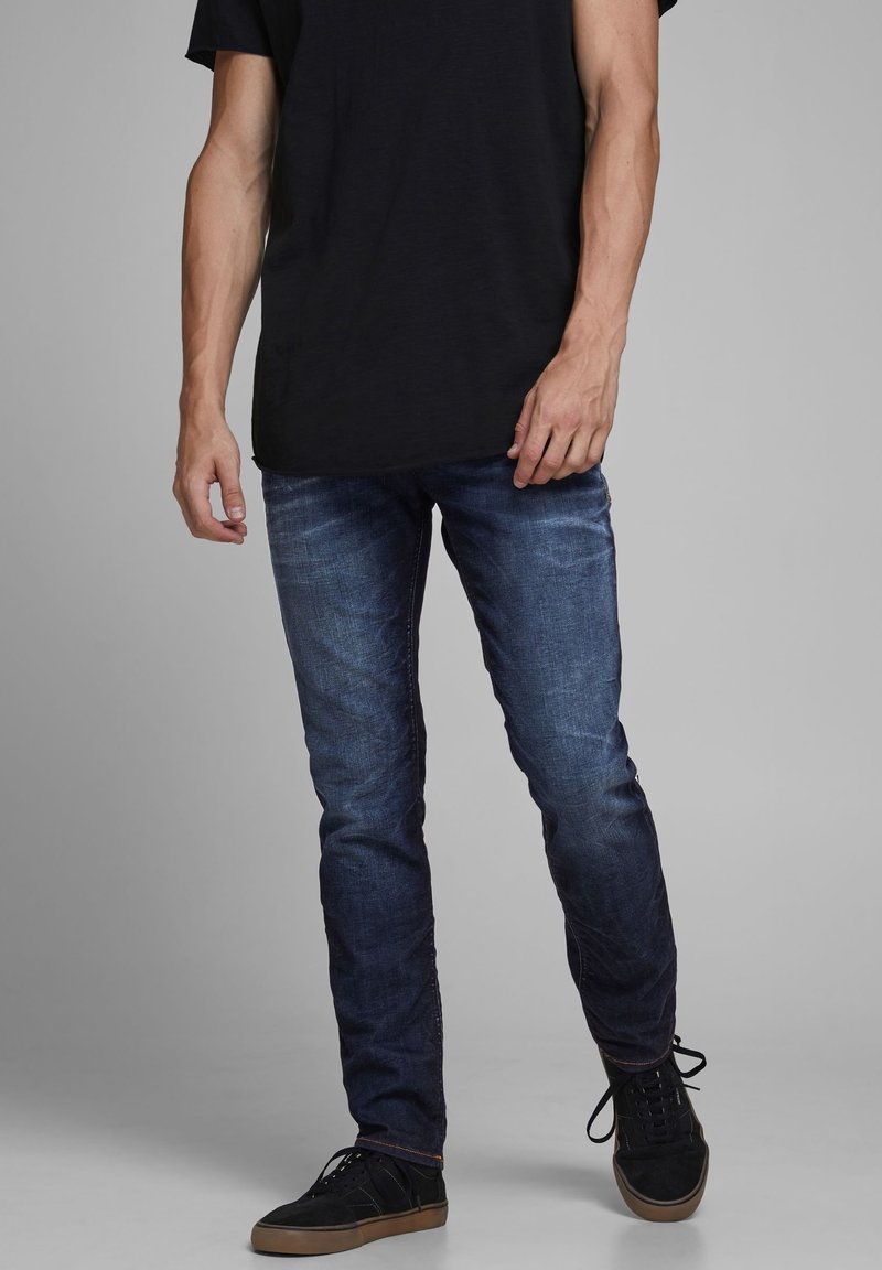 Jeans in denim blu scuro da uomo, con una vestibilità slim, caratterizzati da una leggera sbiaditura. Abbinati a una t-shirt nera a maniche corte e a sneaker nere.