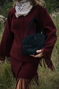 Burgundijas vilnas mētelis ar frizētām piedurknēm un atbilstošām svārkiem; turot nelielu melnu zamša rokas somu ar īsām rokturiem.