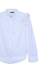 White blouse with a high ruffled collar, decorative button, and ruffled shoulder details. Long sleeves with gathered cuffs. Smooth fabric texture.