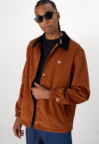 Brown corduroy jacket with black collar, snaps, elastic cuffs, and logo detail. Paired with a black t-shirt and blue jeans.