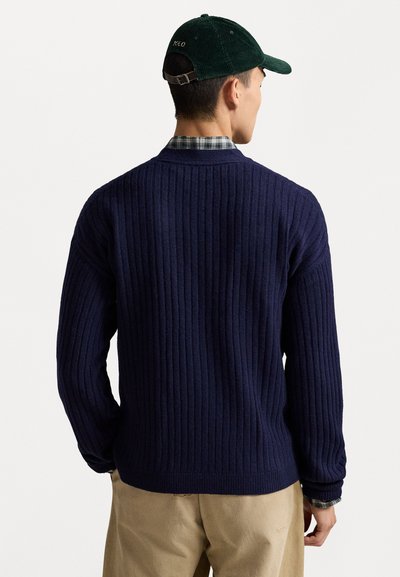 Suéter de punto acanalado en navy, con mangas largas, escote redondo y tejido texturizado. Llevado sobre una camisa de cuadros, combinado con pantalones beige claros.