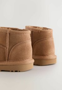 Botine din catifea bej până la gleznă, cu un interior moale, cusături întărite și o talpă din cauciuc texturat. Au o formă rotundă și un profil jos.