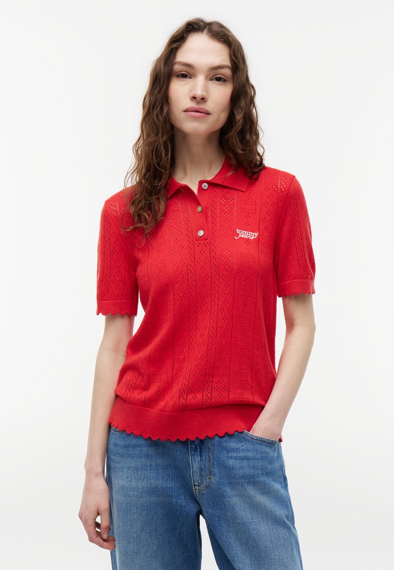 Femme aux cheveux bouclés portant un polo en maille rouge à manches courtes avec des bords festonnés et un jean bleu, debout avec une main dans la poche.