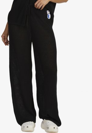 Wide-leg, knitted pants in black with a textured pattern, featuring a drawstring waist and a branded patch on the left side.